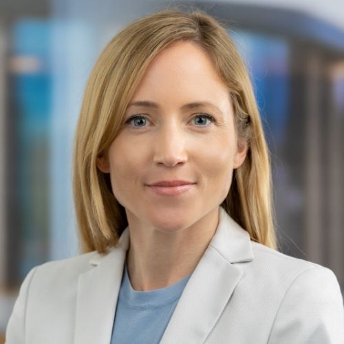 Porträt einer Frau mit blonden Haaren, die in einem hellen Blazer lächelt, vor einem unscharfen Hintergrund.