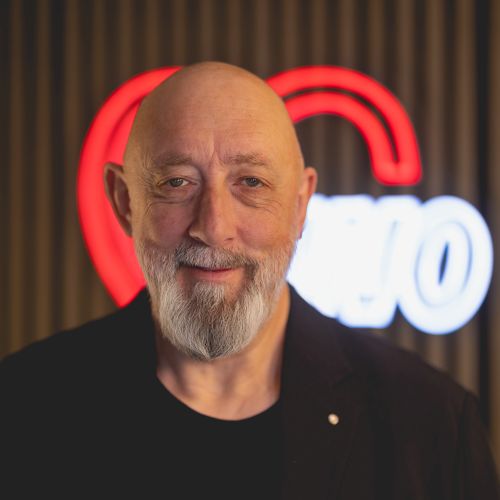 Porträt eines lächelnden Mannes mit Glatze und Bart, vor einem beleuchteten Herz-Logo im Hintergrund.