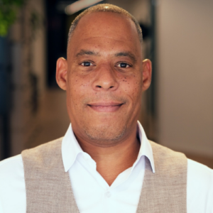 Porträt eines Mannes mit kurzem Haar, lächelnd, in einem hellen Hemd und Weste, vor einem unscharfen Hintergrund.