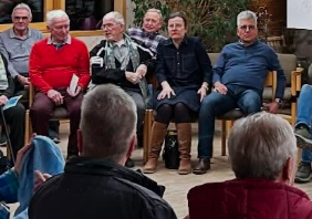 Eine Gruppe von Menschen sitzt in einem Raum, einige sprechen, andere hören zu, mit einem Fokus auf den Redner in der Mitte.