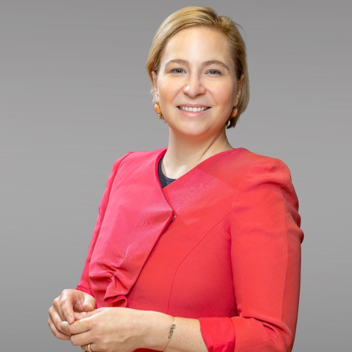 Frau in rotem Oberteil lächelt vor grauem Hintergrund, hält die Hände in einer kommunikativen Pose.