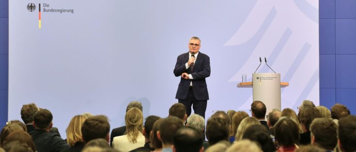 Mann in Anzug spricht vor Publikum in einem offiziellen Raum mit Regierungslogo im Hintergrund.