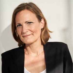 Porträt einer Frau mit schulterlangen, braunen Haaren, die in einem schwarzen Blazer lächelt. Heller Hintergrund.