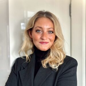 Porträt einer Frau mit blonden, lockigen Haaren, die in einem schwarzen Blazer vor einer hellen Wand steht.