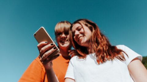 Zwei junge Frauen schauen gemeinsam auf ein Smartphone und lachen, im Hintergrund ist blauer Himmel zu sehen.