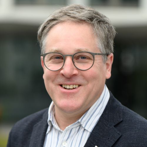 Porträt eines lächelnden Mannes mit Brille und grauen Haaren, im Hintergrund unscharfe Architektur.
