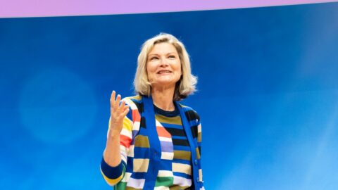 Frau mit blonden Haaren in gestreiftem Pullover spricht lächelnd vor blauem Hintergrund.