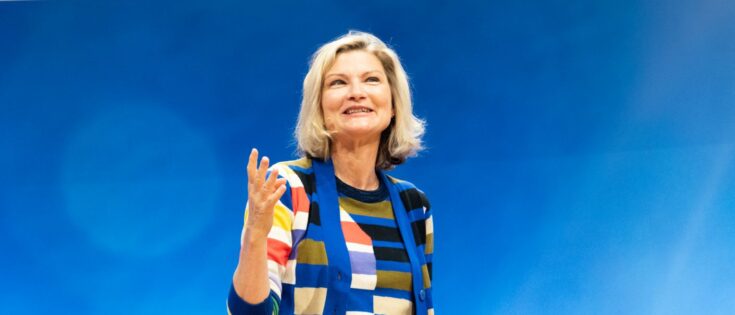 Frau mit blonden Haaren in gestreiftem Pullover spricht lächelnd vor blauem Hintergrund.