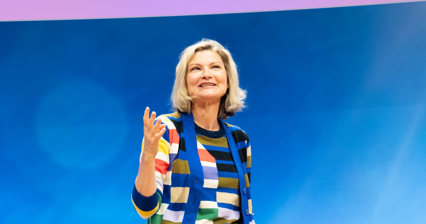 Frau mit blonden Haaren in gestreiftem Pullover spricht lächelnd vor blauem Hintergrund.