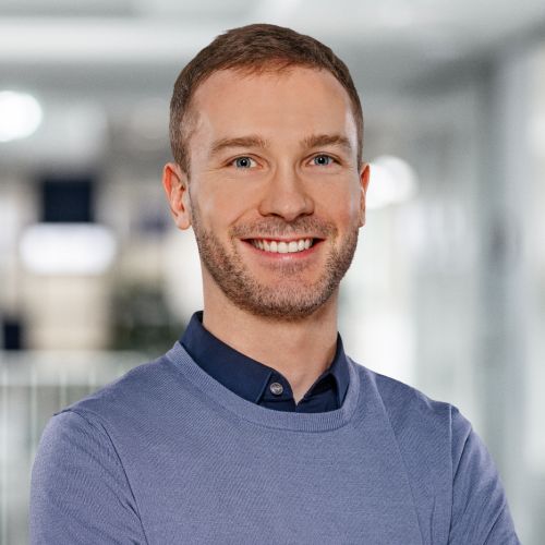 Porträt eines lächelnden Mannes mit kurzem Haar, in einem grauen Pullover und blauem Hemd, vor einem modernen Hintergrund.