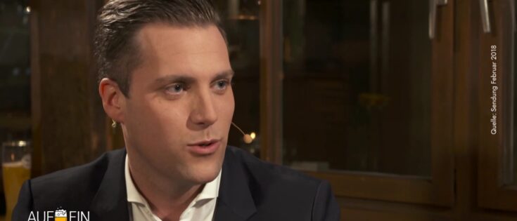 Mann mit kurzen Haaren und Anzug spricht in einem gemütlichen Raum mit Holzdetails.