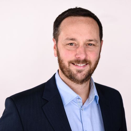 Porträt von Mathias Rauck in Anzug, lächelnd vor neutralem Hintergrund.