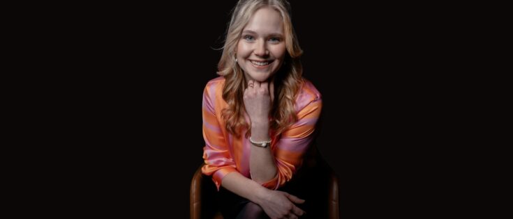 Frau mit blonden Haaren in einem bunten Oberteil sitzt lächelnd auf einem Stuhl vor dunklem Hintergrund.