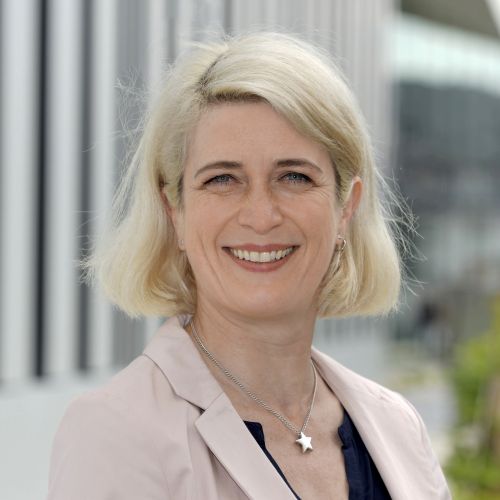 Porträt von Petra Schäfer mit blonden, schulterlangen Haaren, lächelnd vor einem modernen Gebäude.