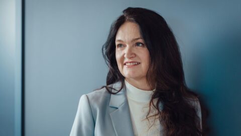 Frau mit langen, dunklen Haaren und lächelndem Gesicht, steht vor einer blauen Wand. Sie trägt einen hellen Blazer.