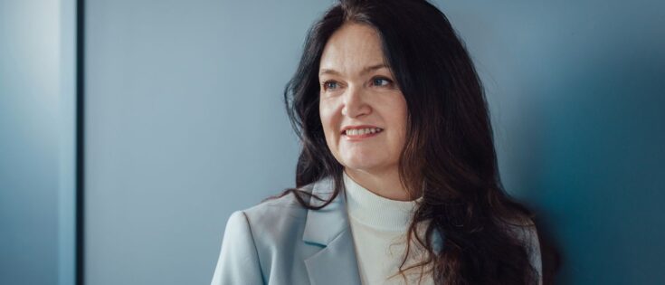 Frau mit langen, dunklen Haaren und lächelndem Gesicht, steht vor einer blauen Wand. Sie trägt einen hellen Blazer.