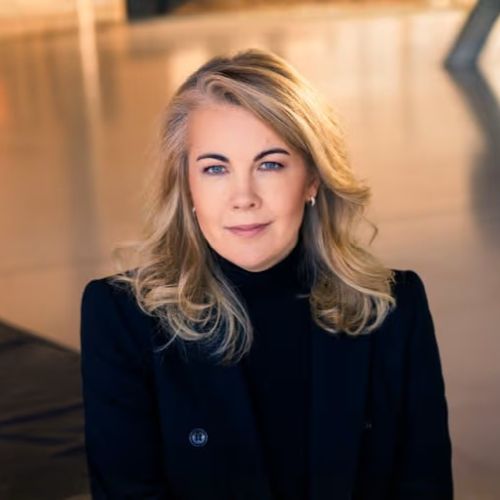 Porträt einer Frau mit blonden, lockigen Haaren, die in einem schwarzen Outfit vor einem modernen Hintergrund sitzt.