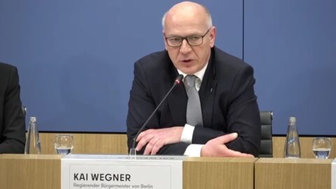 Mann mit Brille und Anzug spricht am Tisch, vor ihm ein Schild mit seinem Namen und Titel.