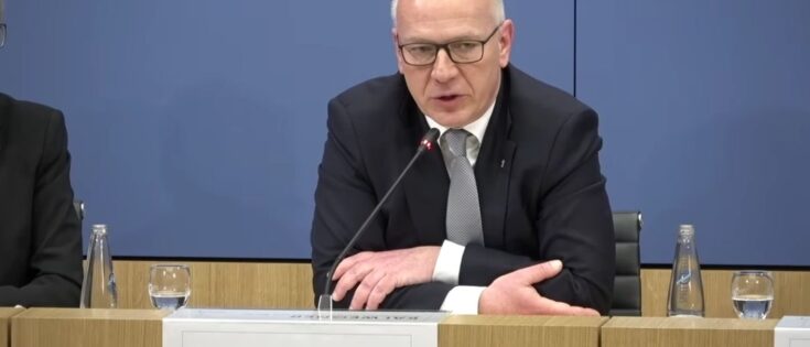 Mann mit Brille und Anzug spricht am Tisch, vor ihm ein Schild mit seinem Namen und Titel.