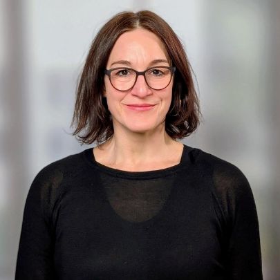 Porträt von Vanessa Zaher mit kurzen, braunen Haaren und Brille, in einem schwarzen Oberteil vor unscharfem Hintergrund.