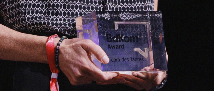 Hände halten einen klaren Preis mit der Aufschrift "BaKom Award - Team des Jahres" auf einer Bühne.