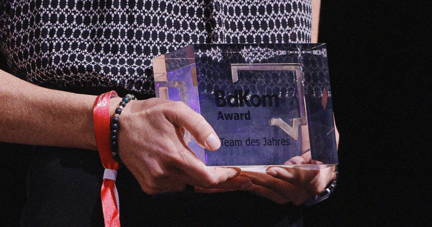 Hände halten einen klaren Preis mit der Aufschrift "BaKom Award - Team des Jahres" auf einer Bühne.