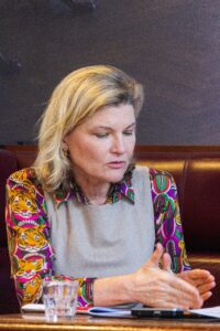 Frau mit blonden Haaren in buntem Oberteil, die konzentriert an einem Tisch sitzt und gestikuliert.