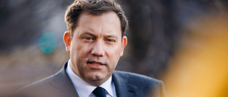 Mann in Anzug spricht in die Kamera, unscharfer Hintergrund mit Lichtpunkten.