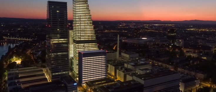Nachtaufnahme der Roche-Gebäude in Basel, beleuchtet vor einem farbenfrohen Sonnenuntergang.