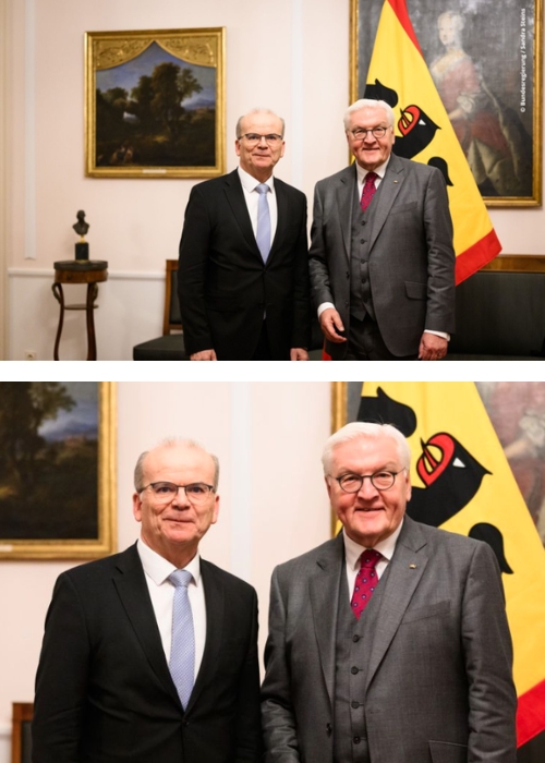 Zwei Männer in Anzügen posieren vor einem Gemälde und einer Flagge, lächelnd für ein Foto.