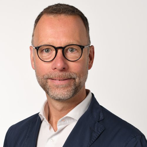 Professional head-and-shoulders portrait of a middle-aged man with round glasses, navy blazer, and white shirt looking at the camera