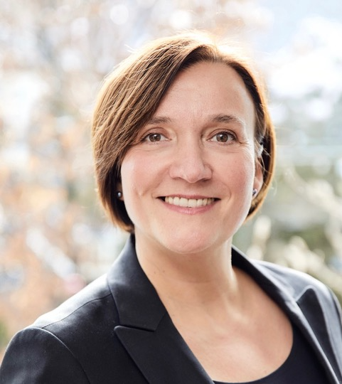 Porträt von Alexandra Appel, lächelnd, mit kurzer brauner Frisur, in einem schwarzen Blazer vor unscharfem Hintergrund.