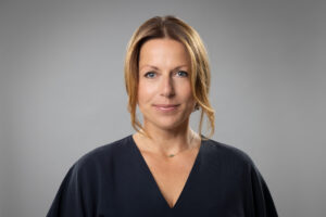 Porträt von Elke Beune mit blonden Haaren, in dunkler Kleidung, vor einem grauen Hintergrund.