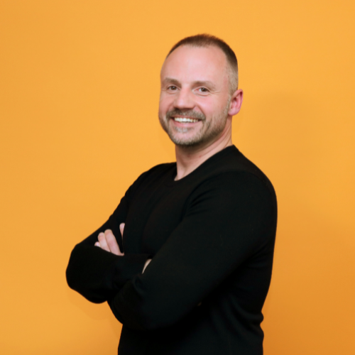 Mann mit kurzem Haar und Bart, lächelt vor orangefarbenem Hintergrund, trägt schwarzes Oberteil, steht mit verschränkte...