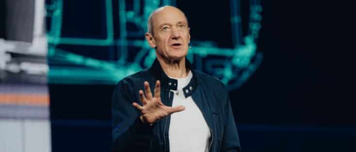 Mann mit Glatze und Jacke spricht auf einer Bühne, im Hintergrund technische Zeichnungen.