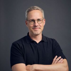 Mann mit Brille und kurzem grauem Haar, trägt ein schwarzes Polo-Shirt, lächelt und steht vor einem dunklen Hintergrund.