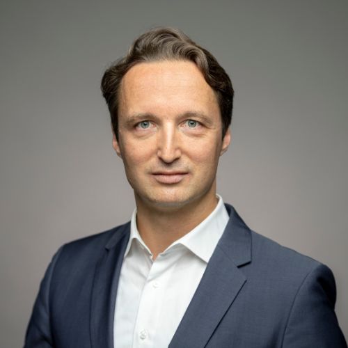Porträt eines Mannes mit braunen Haaren, in einem dunklen Anzug und weißem Hemd, vor einem neutralen Hintergrund.