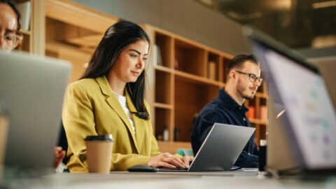 Diverse Teammitglieder arbeiten an Laptops in modernem Büro; eine Frau konzentriert sich auf ihre Aufgaben.