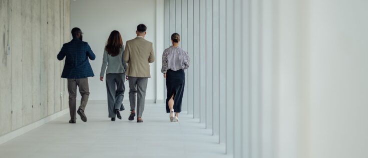 Vier Geschäftspersonen gehen gemeinsam einen modernen Büroflur entlang, umgeben von zeitgenössischer Architektur.