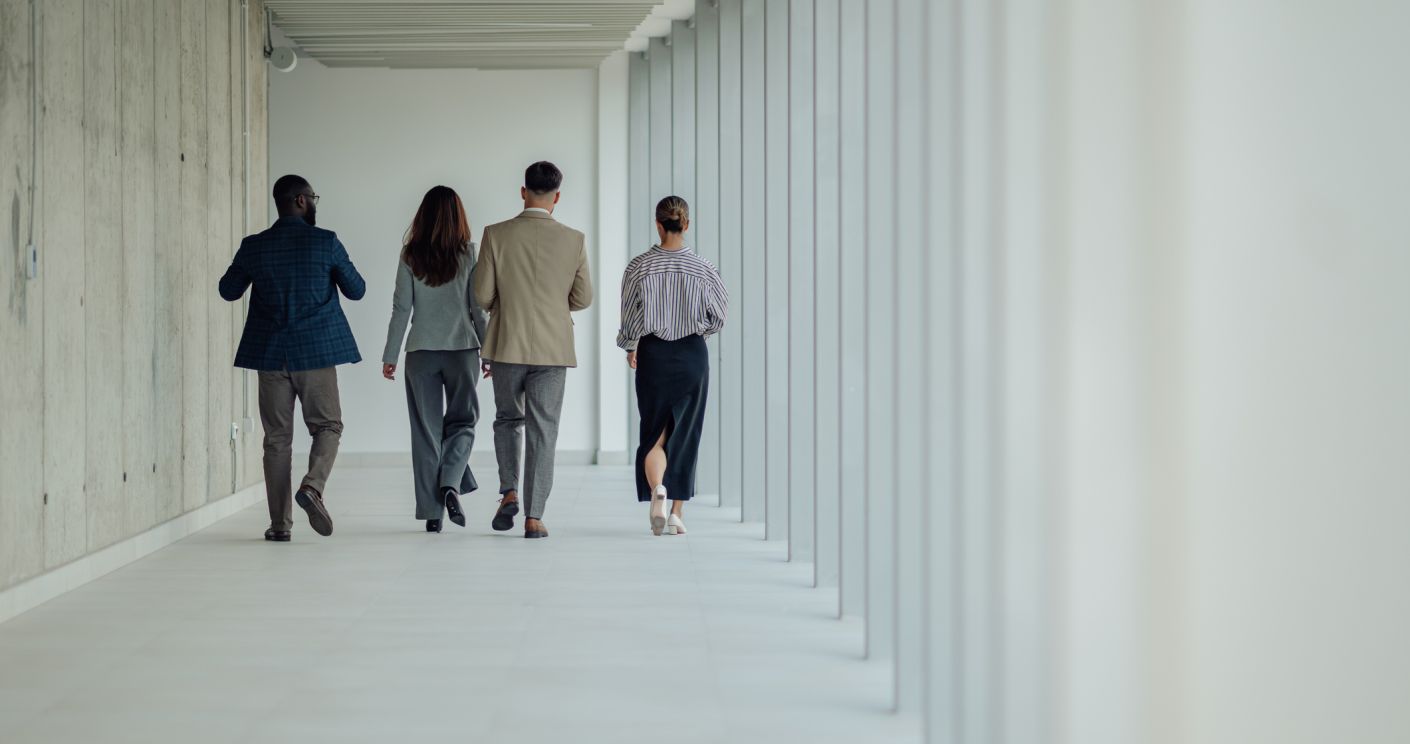 Vier Geschäftspersonen gehen gemeinsam einen modernen Büroflur entlang, umgeben von zeitgenössischer Architektur.