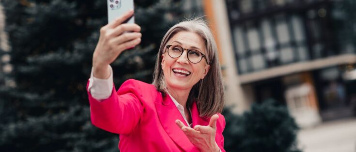 Elegante Seniorin in pinkem Anzug macht draußen ein Selfie und lächelt in einer urbanen Umgebung an einem sonnigen Tag.