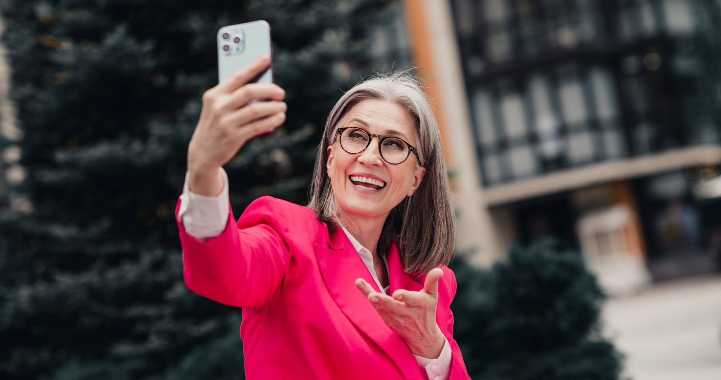 Elegante Seniorin in pinkem Anzug macht draußen ein Selfie und lächelt in einer urbanen Umgebung an einem sonnigen Tag.