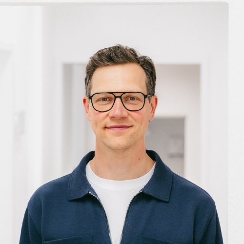 Mann mit Brille und kurzem, dunklem Haar, lächelt vor einem hellen, minimalistischen Hintergrund.