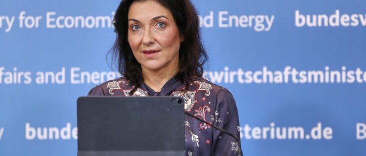 Bundesministerin Katherina Reiche steht vor einem blauen Hintergrund und spricht in ein Mikrofon.