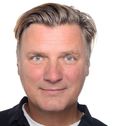 Porträt eines Mannes mit blonden Haaren und blauen Augen, lächelnd vor weißem Hintergrund.