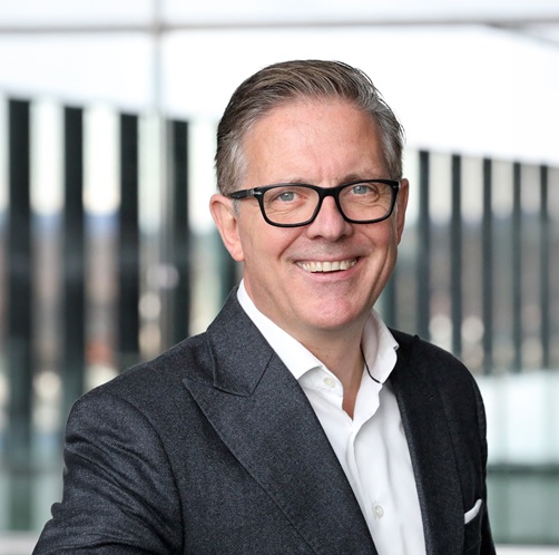 Mann mit Brille und grauem Anzug lächelt vor modernen Fenstern.