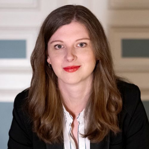 Porträt von Maria Ollesch mit langen, braunen Haaren, blauer Bluse und schwarzem Blazer, vor einem hellen Hintergrund.