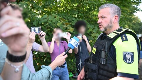 Polizeidirektor gibt Statement an Unfallstelle, während Menschenmenge im Hintergrund mit Handys filmt.