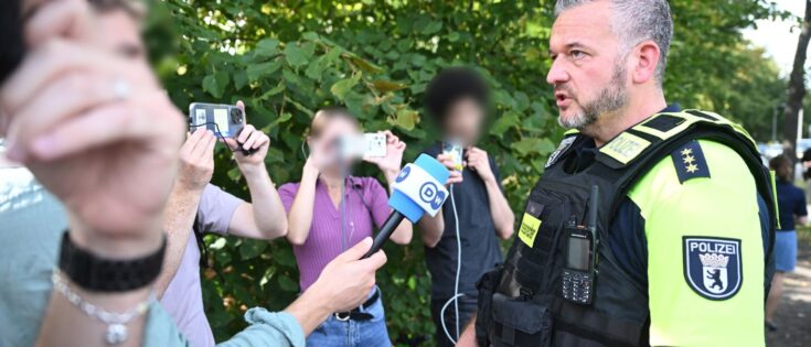 Polizeidirektor gibt Statement an Unfallstelle, während Menschenmenge im Hintergrund mit Handys filmt.