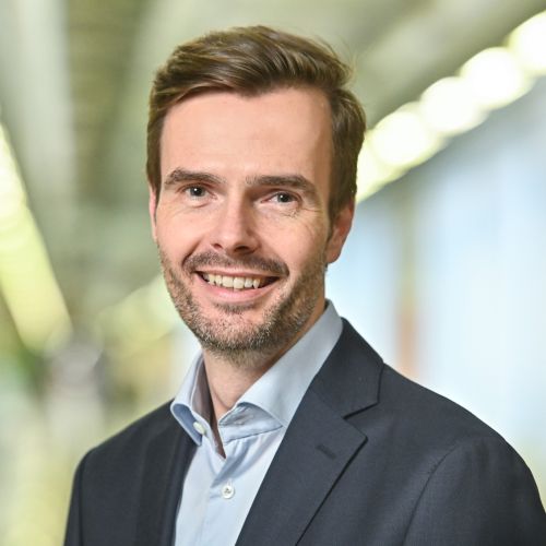 Porträt eines lächelnden Mannes mit kurzem, braunem Haar, in einem Anzug und Hemd, vor einem unscharfen Hintergrund.